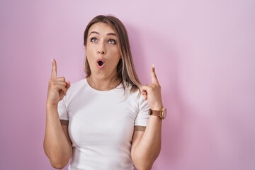 Poster - Blonde caucasian woman standing over pink background amazed and surprised looking up and pointing with fingers and raised arms.