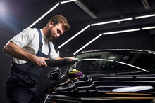 Side view on Young professional car service male worker with orbital polisher, polishing black luxury car hood in auto repair shop, copy space