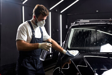 Poster - young man cleaning car with cloth and detergent liquids, car detailing or valeting concept. In auto repair shop, side view on male in protective mask, gloves and uniform