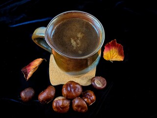 Cup of coffee with autumn leaves and chestnuts against black background