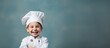 A young boy wearing a chef s hat and uniform standing against a grey background exemplifies the joyous aspect of the culinary profession