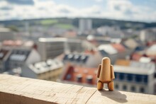 A Wooden Figure Stands On A Ledge Overlooking A City. Blurred City, Urban Background.