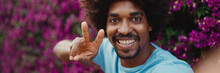 Close-up Portrait Of Young African American Man Shooting Selfie Use Smartphone Stands On Background Wall Covered With Purple Flowers And Smiling. Lifestyle Concept.