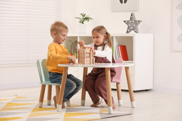 Wall Mural - Little boy and girl playing with wooden house at white table indoors. Children's toys