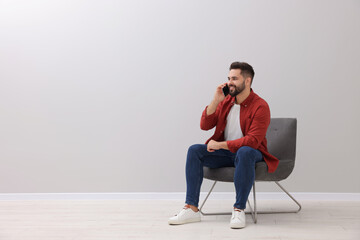 Poster - Handsome man talking on smartphone while sitting in armchair near light grey wall indoors, space for text