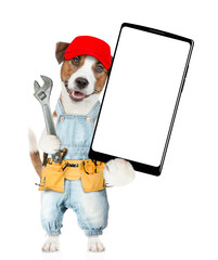 Wall Mural - Funny Jack russell terrier puppy wearing red cap and denim overalls with tool belt showing big smartphone with white blank screen in it paw. Isolated on white background