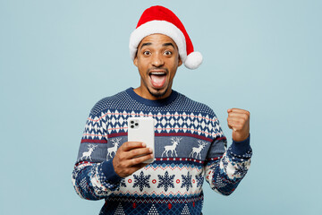 Young man wear knitted sweater Santa hat posing hold in hand use mobile cell phone do winner gesture isolated on plain pastel blue background Happy New Year 2024 celebration Christmas holiday concept