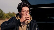 Close-up shot of a guy sitting in the trunk of an SUV in the steppe and drinking tea from thermos while admiring nature