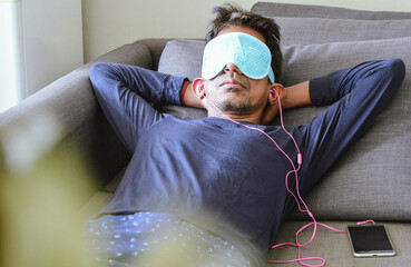 A South Asian man relaxing wearing a heated reusable eye mask and listening to music on the sofa. Selective focus.