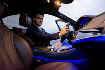 Wall Mural - Businessman sitting on drivers seat of luxury sport car at dusk