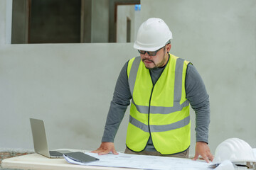 Asian architect Checking construction plans from blueprints and on a laptop. Plans instructing workers. Men wearing green reflective vests and hard hats. At a work desk on a construction site