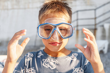 Handsome smiling boy with diving mask on head rests on beach closeup. Little child with scuba equipment for underwater swimming at resort