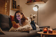 © olezzo - A young woman spends a cozy winter evening at home lying on the sofa looking at her smartphone. Winter holidays, Christmas and online surfing concept