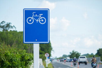 Blue bicycle lane sign alongside a busy road with passing vehicles and greenery under a clear sky, highlighting transportation and cycling themes