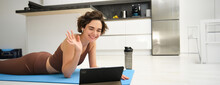 Smiling Brunette Woman In Sportswear, Waves Hand, Says Hi At Tablet, Records Video Sports Blog, Fitness Training Session Online, Instructor Shows Workout Elements, Uses Rubber Mat At Home In Kitchen