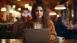 A young woman is seen working on a laptop at a cafe table, looking directly into the camera, She appears to be a freelancer or student,