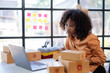 © amnaj - Young African American woman online selling. Startup small business entrepreneur or freelance african woman using a laptop with box. Online marketing packaging box and delivery, SME concept.