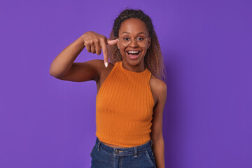 Wall Mural - Young happy African American woman enthusiastically looks at screen and points finger down recommending that you pay attention to news or great offer from supermarket stands in purple studio.