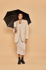 Wall Mural - happy african american woman in midi dress and autumnal blazer standing under umbrella on beige