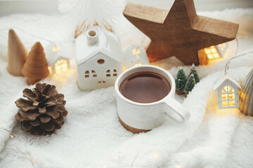 Wall Mural - Stylish cup of tea with modern christmas houses, pine cone, wooden star and tree decor, golden lights on warm blanket on windowsill. Cozy Christmas, scandinavian style. Christmas background