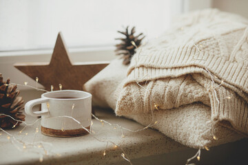 Wall Mural - Stylish cup of tea with cozy knitted sweater, pine cone, wooden star and golden lights on windowsill in festive scandinavian room. Cozy winter still life. Autumn hygge