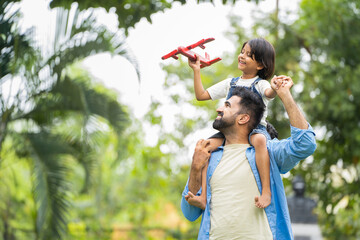 happy smiling indian girl kid playing with airplane toy by sitting on father shoulder at park - conc