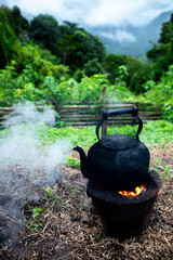 Black metal kettle on burning bonfire with hot flames at campsite against  nature campsite and mountains are in the background.