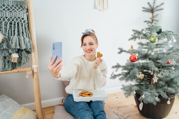 Happy Young Woman in cozy sweater taking selfie photo while relaxing on floor cushions near potted christmas tree in modern Scandi interior home. Cozy winter holiday. Content creating. Selective focus