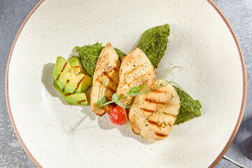 Canvas Print - Close-up top view of grilled chicken fillet, avocado, and spinach on a ceramic plate. The dish stands out against a grey concrete background with hard shadows, making it a healthy culinary choice