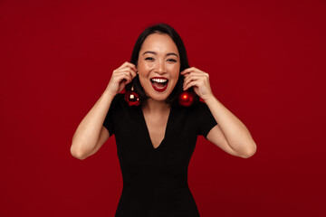 Cheerful asian woman holding Christmas tree toys like earrings while standing isolated over red wall
