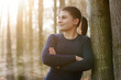 Athletic young sporty woman after workout, running and exercises leaning relaxed on a tree on a sunny morning in winter or autumn in forest	