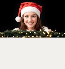 Happy Young Woman In Xmas Santa Hat Pose Behind Empty White Board With Copy Space Decored With Christmas Branch And Lights Isolated On Black 
