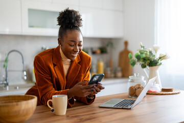 Wall Mural - Charming African businesswoman texting over smart phone while working over laptop at desk in home office.