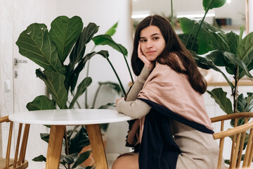 Young pretty woman sitting inside cafe waiting for order or boyfriend, relaxing smiling happily, joyful posing enjoying atmosphere near plant flowerpot

