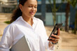 © Taras Grebinets - Closeup portrait of a charming adult woman checking new mobile app on smartphone, standing outdoor with laptop in hands