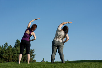 Two middle-aged women doing fitness in city park, bending body to side, blue sky background, back view. Healthy lifestyle, yoga, Pilates, weight loss. overweight woman doing yoga with trainer.