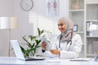 © Liubomir - Portrait Muslim female doctor in hijab, woman doctor with tablet computer using application, smiling satisfied work working inside medical office, in white medical coat, sitting at table with laptop