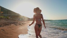 Happy Young Girl In A Hat And Swimsuit Runs Along The Beach At Sunset. Holiday At Sea. Beautiful Sunset Light On The Coast.