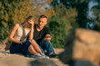 A young man quarrels with his girlfriend who turns away from him offended while sitting on sandy bank of the river