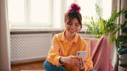 Wall Mural - Young Asian woman with bright pink locks of hair talking on the phone while sitting on the armchair by the window in the living room at home.