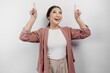 Excited Asian woman employee wearing cardigan pointing at the copy space above her, isolated by white background