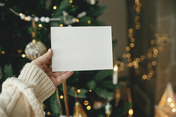 Christmas card mock up. Hand holding empty greeting card on background of stylish decorated christmas tree with golden lights. Space for text. Season greetings template and vintage ornaments