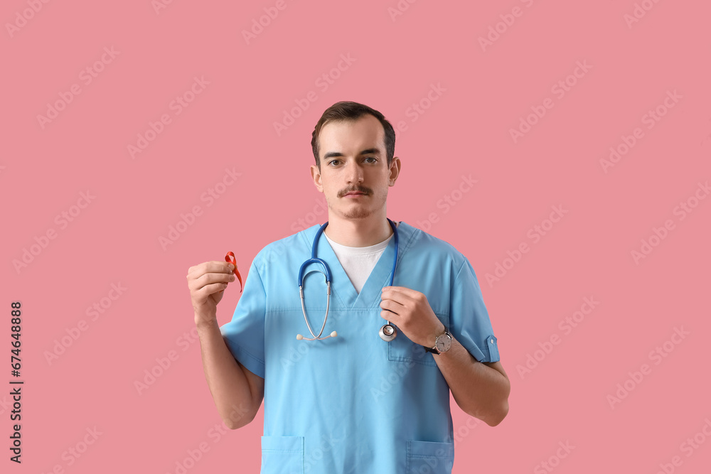 Young male doctor with red ribbon on pink background. World AIDS Day concept
