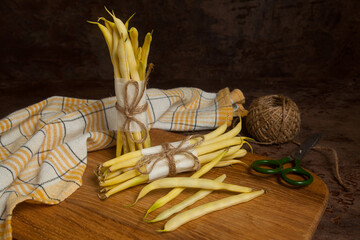 Cutting board with two bunch and several pods of flat runner bean pods heap with yellow kitchen towel, scissors and ball of thread on wooden background. .