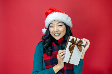 Wall Mural - Pretty smiling asian woman in warm christmas sweater and santa hat holding gift box as present with red background for season celebration