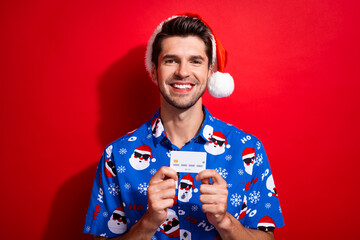 Wall Mural - Portrait of positive cheerful person dressed santa hat blue shirt presenting debit card in hands isolated on vibrant red color background