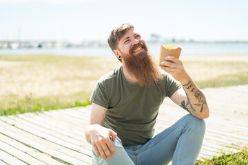 Wall Mural - Redhead man with beard holding fried chips at outdoors looking up while smiling