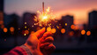 A hand holding a brightly lit sparkler at dusk, with a backdrop of warm bokeh lights from a city street.