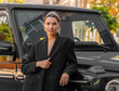 beautiful woman in black office suit leans against black crossover car outside, urban city background