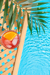 Wall Mural - Glass of cold Negroni cocktail and palm leaf on wooden grid near swimming pool